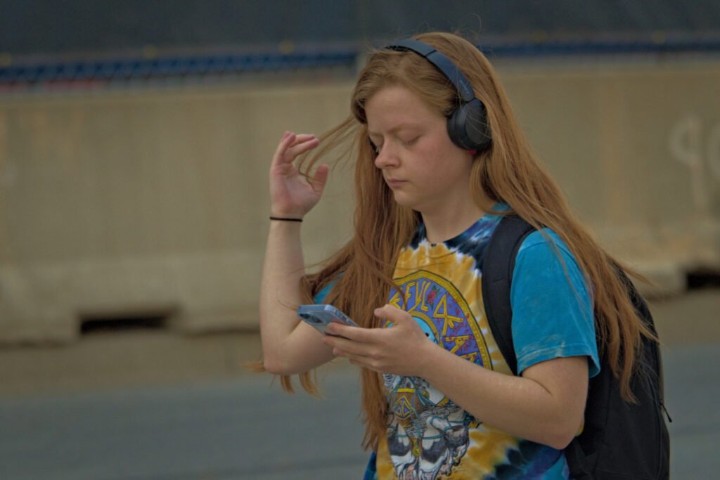 West Lafayette Downtown (Chauncey Village), 2026-03-29 - Woman Playing with Her Hair While Using Her Cellphone and Wearing Headphones